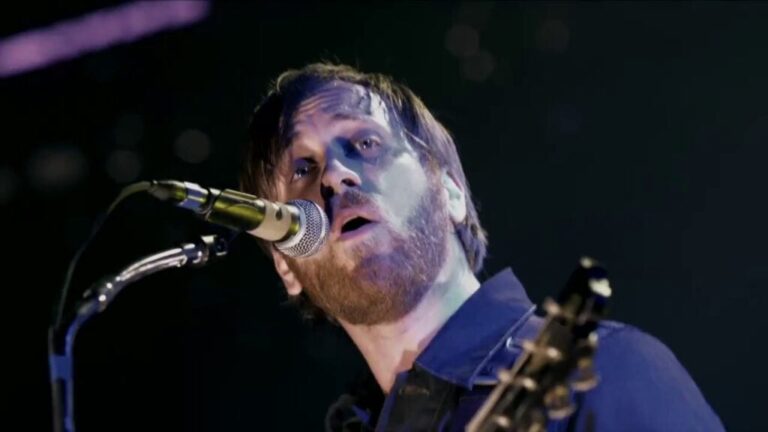 Singer performing live on stage, singing into a microphone under concert lighting.