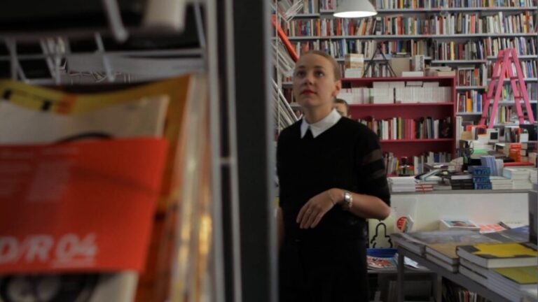 Woman standing in a bookshop interior surrounded by shelves of books.