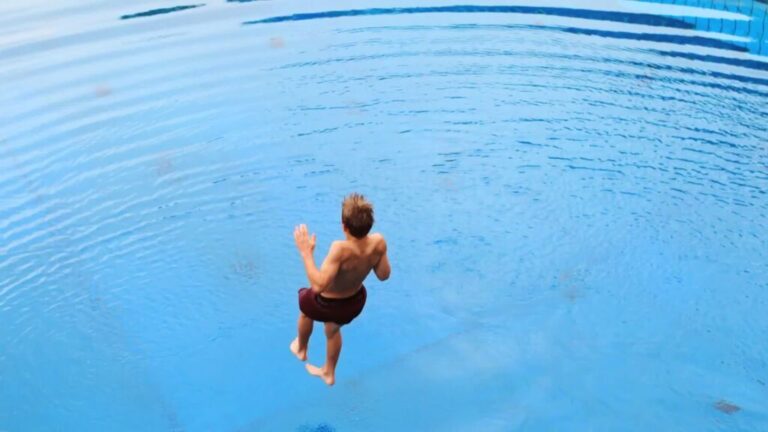 Young man jumping into a bright blue swimming pool, captured mid-air from above.