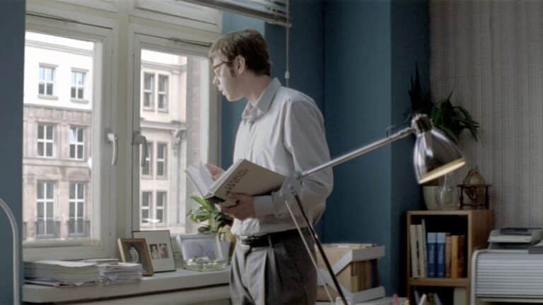 Man standing by a window in a quiet interior, holding a book and reflecting.