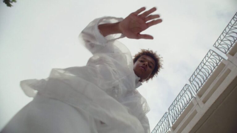 Person in flowing white clothing seen from a low angle, reaching upward against the sky.