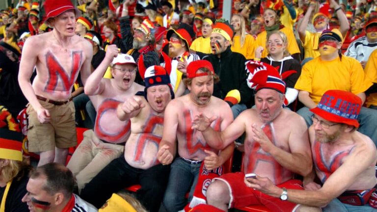 Group of football fans with painted torsos and hats cheering loudly in a packed stadium.