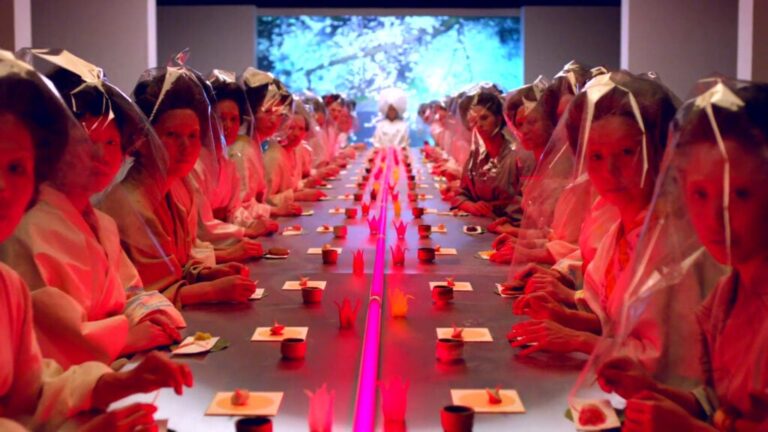 Rows of women in translucent garments sit facing each other at a long table under red lighting.