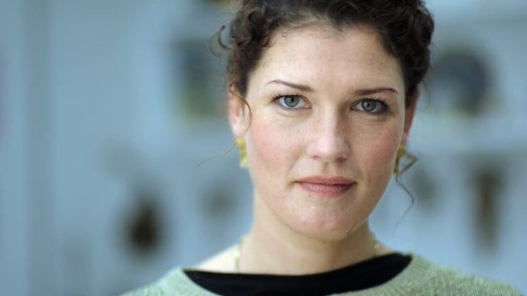 Close-up portrait of a woman with curly hair and blue eyes, looking directly at the camera against a softly blurred background.