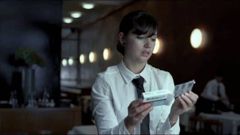A woman in a restaurant uniform examines a small packet while standing in a softly lit interior.
