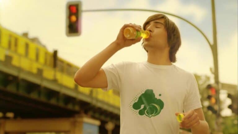 A young man drinks from a small bottle while standing at a city intersection in daylight.