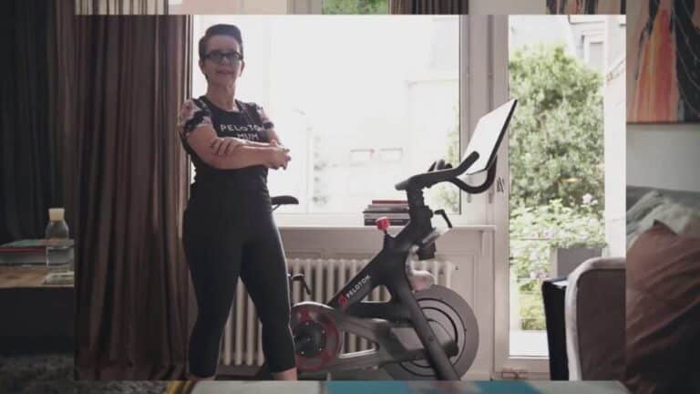 A woman stands beside a Peloton bike in a bright living room with arms crossed.