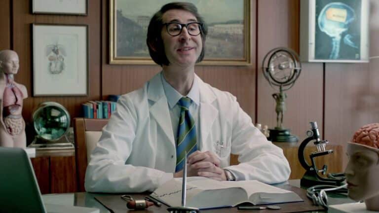 A scientist in a white lab coat sits at a desk with a microscope and anatomical models, smiling as he speaks in a laboratory office.