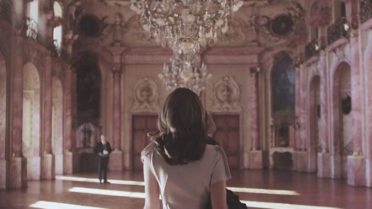 Woman standing in an ornate historic hall beneath a chandelier, viewed from behind.